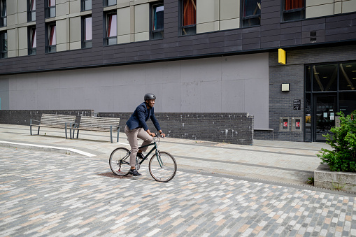 A mature black man wearing formal businesswear and a helmet on a summers day in a city. He is riding bicycle as he sustainably commutes to work.