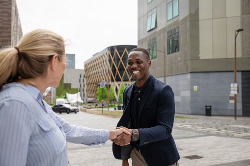 A mature black man wearing formal businesswear being greeted by his new employer, a mature caucasian woman on a summers day in a city. They are shaking hands and greeting each other positively.