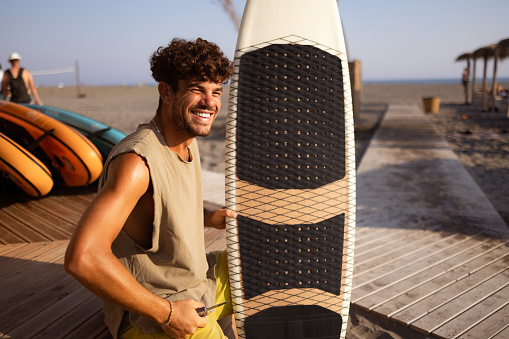 Young Caucasian man enjoying his vacation, sitting on a beach with a surfboard and smiling