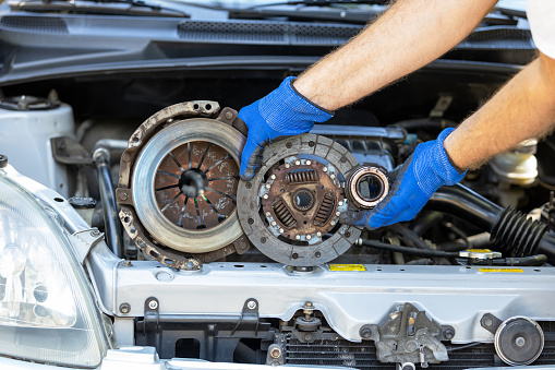 Automotive technician holding used car pressure plate, clutch disc and release bearing in front of the vehicle engine