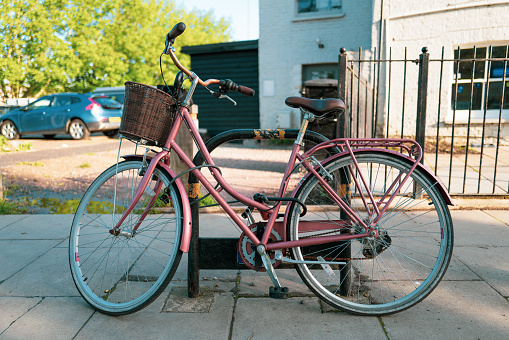 Old fashioned pink bicycle with wicker basket locked to a bike stand on a city street in London, UK.