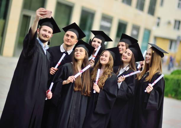 group of graduate students of masters in black robes and square hats take selfies on a smartphone camera a group of graduate students of masters in black robes or mantle and square hats with diplomas in their hands take selfies on a smartphone camera... Master’s Degree stock pictures, royalty-free photos & images