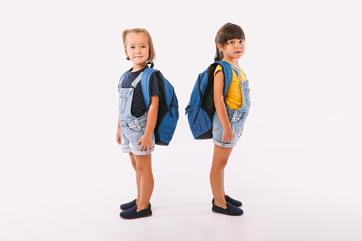 Two little girls, one with blonde hair and the other with black hair, dressed in denim blue overalls, with a backpack, ready for back to school, sideways, on white background.