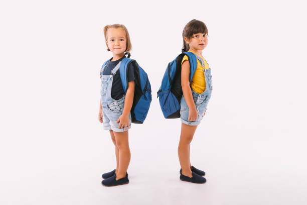 Two little girls, one with blonde hair and the other with black hair, dressed in denim blue overalls, with a backpack, ready for back to school, sideways, on white background. stock photo