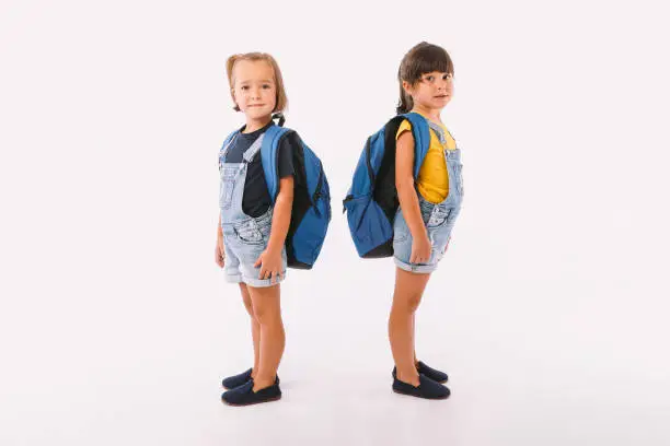 Photo of Two little girls, one with blonde hair and the other with black hair, dressed in denim blue overalls, with a backpack, ready for back to school, sideways, on white background.