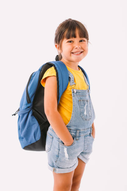 bambina con i capelli neri vestita con una tuta di jeans e una t-shirt blu, con uno zaino pronto per tornare a scuola, al suo fianco, su sfondo bianco - schoolgirl school children isolated child foto e immagini stock