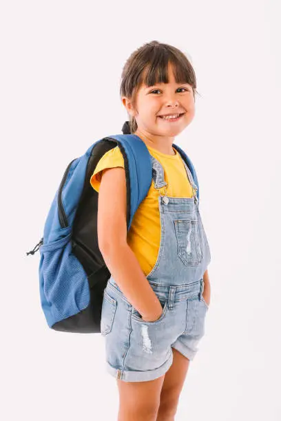 Photo of Little girl with black hair dressed in a denim overalls and a blue t-shirt, with a backpack ready for going back to school, on her side, on white background