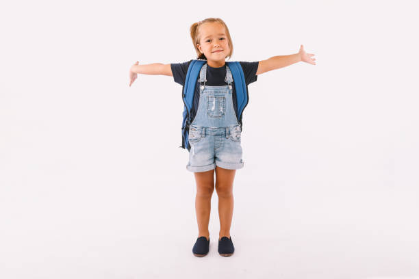 Little girl with blond hair dressed in a denim overalls and a blue shirt, with a backpack ready for going back to school, with her arms open, very happy, on white background stock photo
