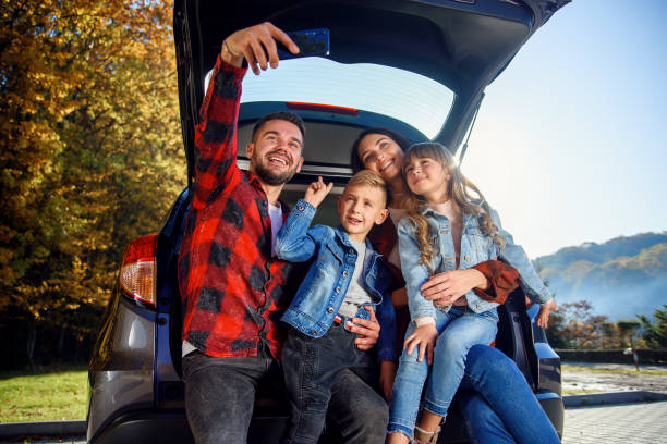 szczęśliwi stylowi rodzice ze swoimi uroczymi dziećmi robią zabawne selfie na smartfonie, siedząc w bagażniku. szczęśliwa nowoczesna koncepcja rodzinna. - family trip zdjęcia i obrazy z banku zdjęć