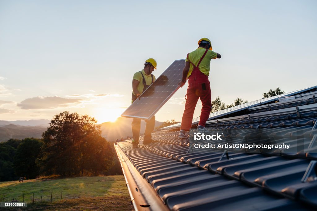 Workers Installing Solar Panels On Wooden House In Nature At Sunset. Shot Of Professional Workers Installing Solar Panels On A Roof At Sunset. Sustainable Energy Stock Photo
