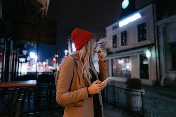 Photo of a young woman drinking hot drink, using smart phone and walking through the town during the Christmas holidays