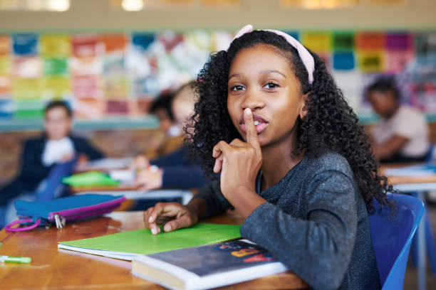 photo d’une jeune fille assise dans sa classe d’école avec son doigt sur ses lèvres dans un geste de silence - african descent confidence african culture education photos et images de collection