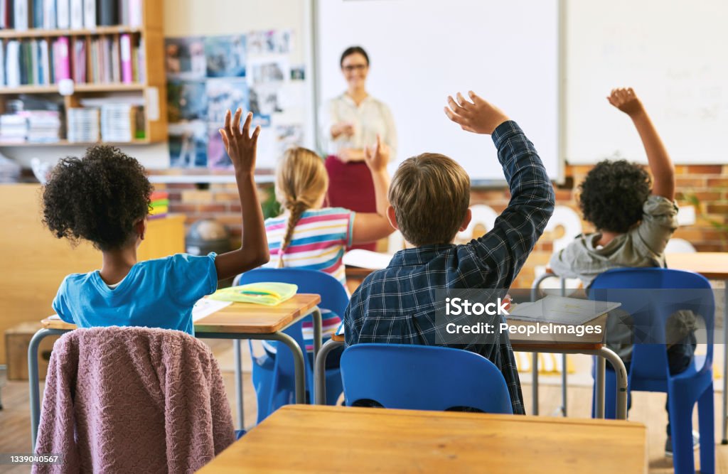 Shot of an unrecognizable group of children sitting in their school classroom and raising their hands to answer a question Who shall we choose? Classroom Stock Photo