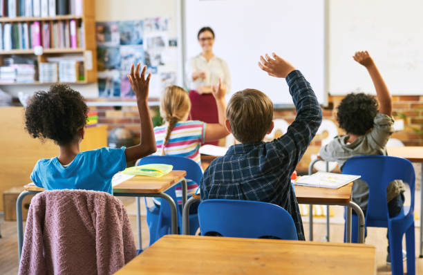 снимок неузнаваемой группы детей, сидящих в школьном классе и поднимающих руки, чтобы ответить на вопрос - учёный стоковые фото и изображения