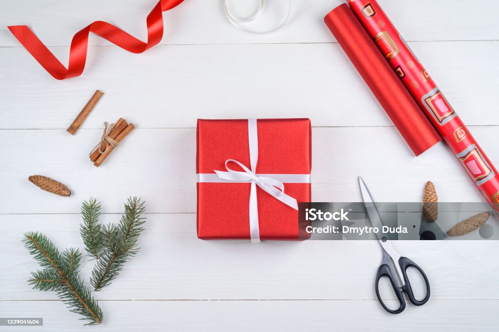 Top view of red Christmas gift box, wrapping paper, ribbon, fir tree branches, cones, cinnamon and scissors on white wooden background, copy space. Gift wrapping, packing.  Flat lay, free space Wrapping Paper Stock Photo