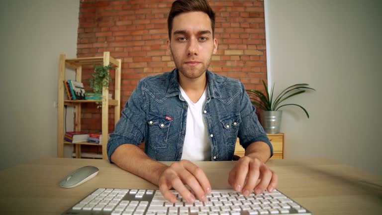 POV of focused handsome male freelancer working at desktop computer, typing on wireless keyboard and using mouse, looking at camera.