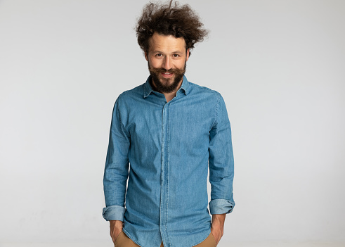happy young man in denim shirt smiling and holding hands in pockets while standing and posing against gray background in studio, portrait