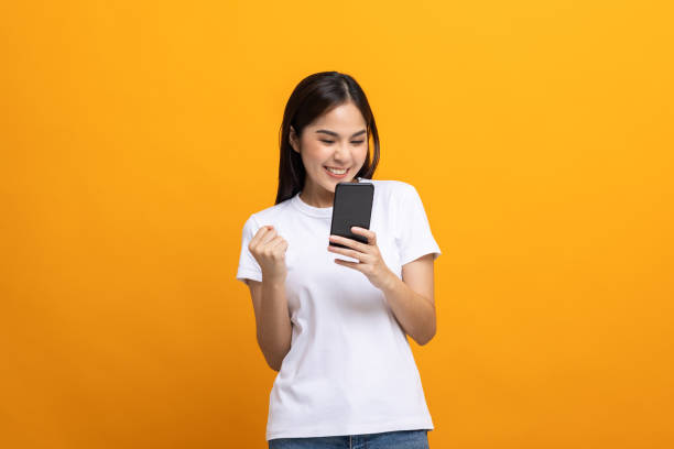 istock Pretty young asian woman using smartphone standing on isolated yellow background feeling happy. Shopping online payment with mobile phone. Excited and surprised female holding cellphone. 1339053261