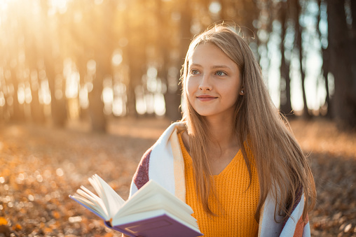 Smiling blond girl likes to read books outdoors in autumn park