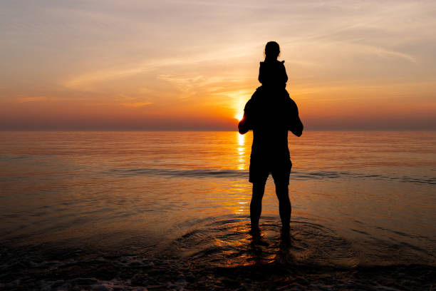 Man with daughter on shoulders silhouet in the calm ocean sea watching the sunset. A father with his daughter on his shoulders (silhouet) are standing in the calm ocean sea watching the sunset in the Netherlands. indian summer stock pictures, royalty-free photos & images