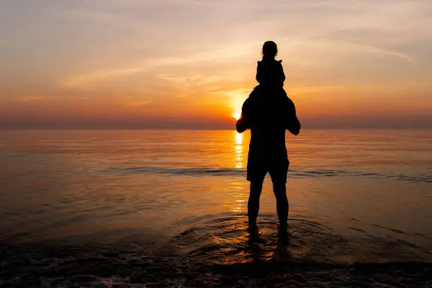 A father with his daughter on his shoulders (silhouet) are standing in the calm ocean sea watching the sunset in the Netherlands.