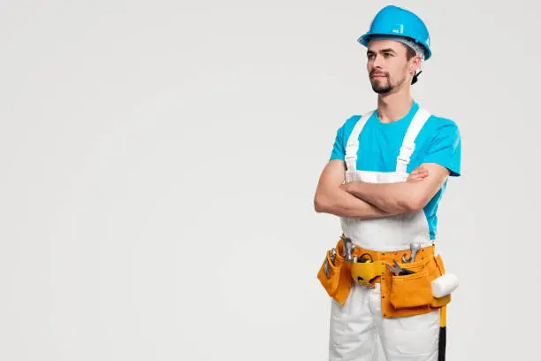 Serious young bearded male worker in overall and hardhat with professional tools standing with crossed arms and looking away with confidence against white background