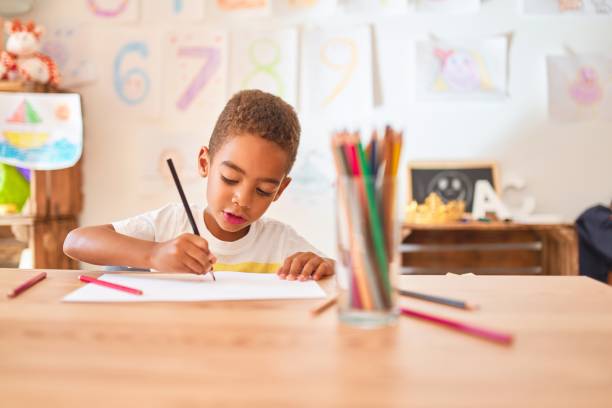 beau bambin afro-américain assis à dessiner en utilisant du papier et des crayons sur le bureau à la maternelle - little boys preschooler child learning photos et images de collection