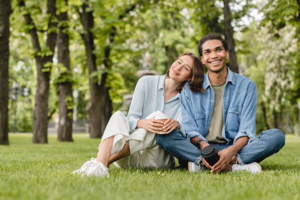 jovens estudantes de casal mestiços e namoradas passando um tempo juntos em encontros românticos sentados na grama verde do gramado no parque da cidade andando ao ar livre. conceito de amor e relacionamento - boyfriend heterosexual couple married activity - fotografias e filmes do acervo