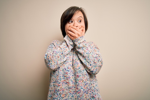 Young down syndrome woman standing over isolated background shocked covering mouth with hands for mistake. Secret concept.