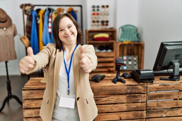 junge frau mit down-syndrom, die als managerin in einer einzelhandelsboutique arbeitet, die positive geste mit der hand genehmigt, lächelt und freut sich über den erfolg. gewinnergeste. - thumbs up human thumb human finger brown hair stock-fotos und bilder