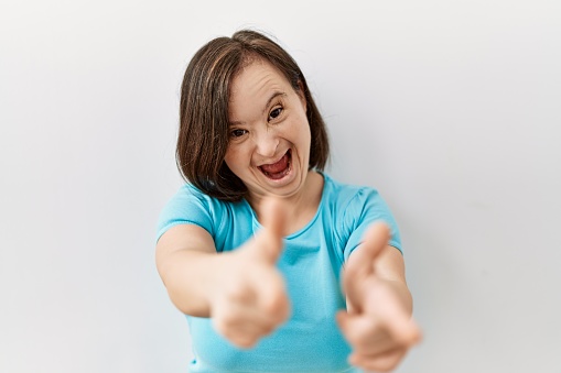 Young down syndrome woman standing over isolated background pointing fingers to camera with happy and funny face. good energy and vibes.