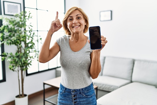 Middle age blonde woman working holding smartphone showing screen surprised with an idea or question pointing finger with happy face, number one