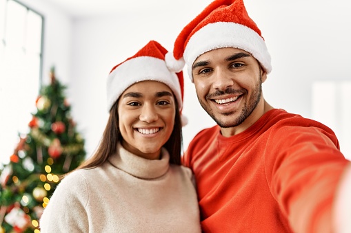 Young hispanic couple wearing christmas hat making selfie by the smartphone at home.