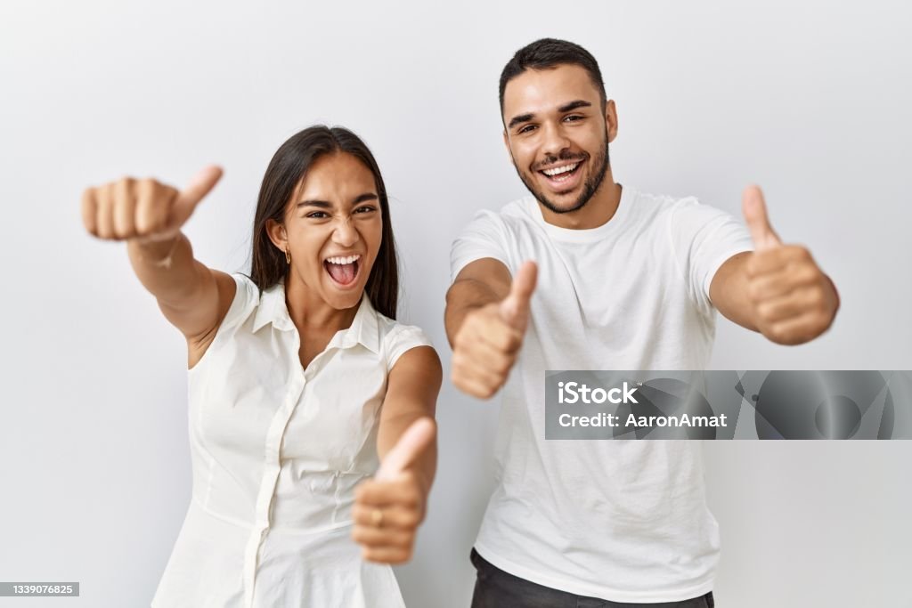 Young interracial couple standing together in love over isolated background approving doing positive gesture with hand, thumbs up smiling and happy for success. winner gesture. Couple - Relationship Stock Photo