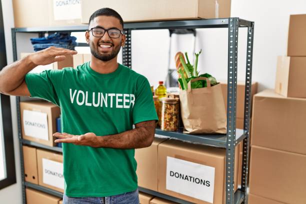 istock Young indian man volunteer holding donations box gesturing with hands showing big and large size sign, measure symbol. smiling looking at the camera. measuring concept. 1339077937