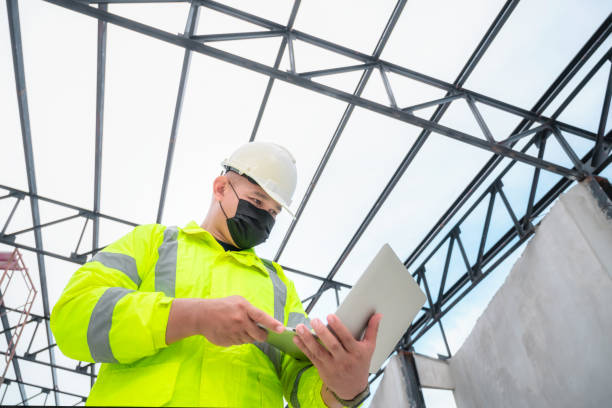istock Civil engineer wearing mask inspect construction site structure and plans in laptop. Civil engineer inspects the actual construction site.  Civil engineer holds laptop to inspect the building. 1339085177