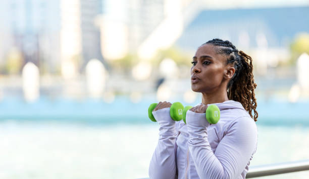A mature African-American woman in her 40s exercising outdoors, in a city park on the waterfront, by a large fountain. She is working out with hand weights.