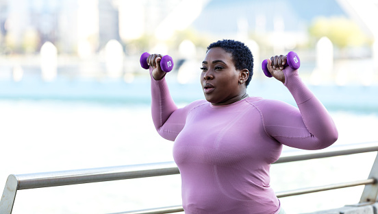 A mid adult African-American woman in her 30s exercising in a city park on the waterfront. She is a plus size model with a large build. She is wearing long sleeved sports clothing, working out with hand weights.