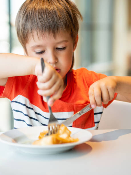 istock Cute kid in red t-shirt eats pancakes with knife and fork. Curious boy with funny expression on face. Tasty pastry for breakfast. 1339088440