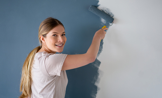 Happy woman painting a wall of her house using a paint roller and looking at the camera smiling - home improvement concepts