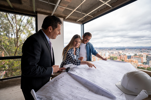 Happy Latin American real estate developer showing blueprints of a house to a couple at a construction site - home ownership concepts