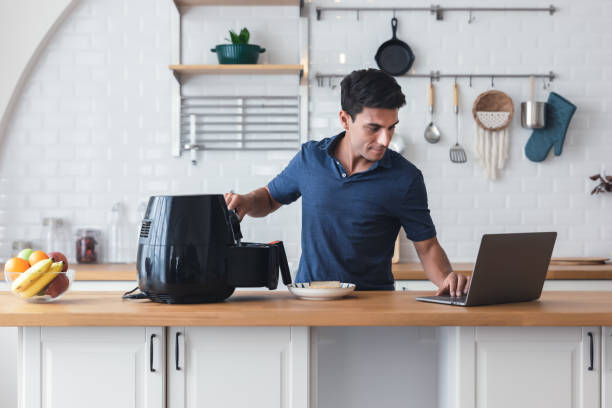 Happy young man standing at kitchen table and cooking toast bread by Air Fryer machine, watching culinary TV show online on laptop. New normal work lifestyle at home. Technology smart cooking device. joven feliz de pie en la mesa de la cocina y cocinando pan tostado por la máquina air fryer, viendo un programa de televisión culinario en línea en una computadora portátil. nuevo estilo de vida normal de trabajo en casa. dispositivo de cocina intelige