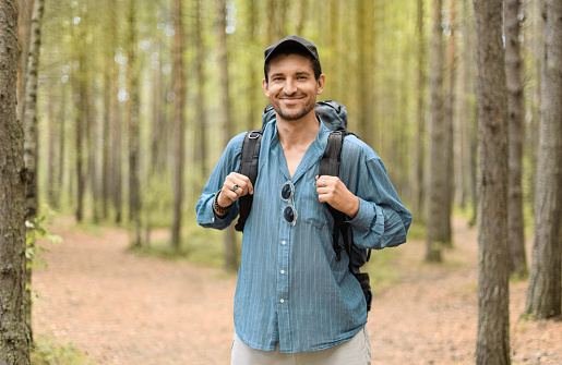 Smiling man, 40 years old, with his tourist backpack is standing in a forest.