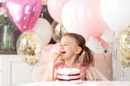 Birthday party of little princess in festive dress. Child celebrates holiday. She sits at table over balloons background and eat birthday cake use hands.Empty space for text.Portrait birthday kid