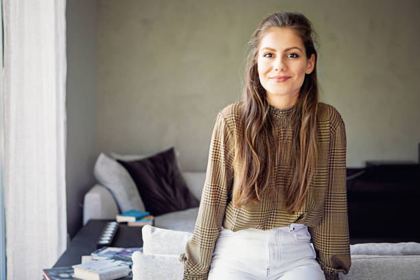 porträt einer jungen frau, die durch fenster in einem wohnzimmer schaut - living room people joy happiness stock-fotos und bilder