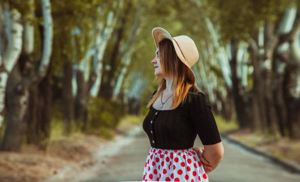 a girl in a summer hat in a skirt with polka dots stands with her eyes closed against the background of an arch of birches and enjoys the fresh air. - garment fragility women skirt imagens e fotografias de stock