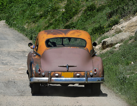 Rear view of a old rusty car driving down a country road.