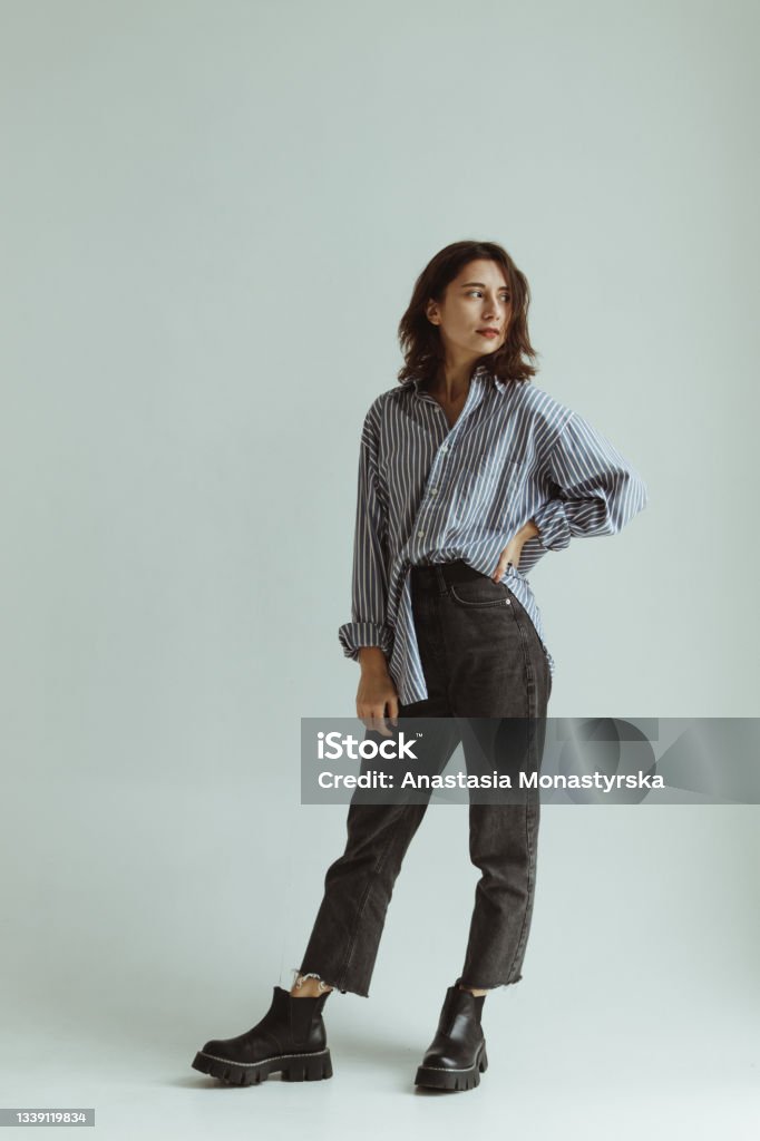 A girl  in a man's loose shirt, chelsea boots and jeans stands in a white studio. Women Stock Photo