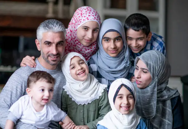 A beautiful family portrait of a Muslim family at home. There are six children in the family. They are all seated close to each other in their living room. They are all smiling at the camera.