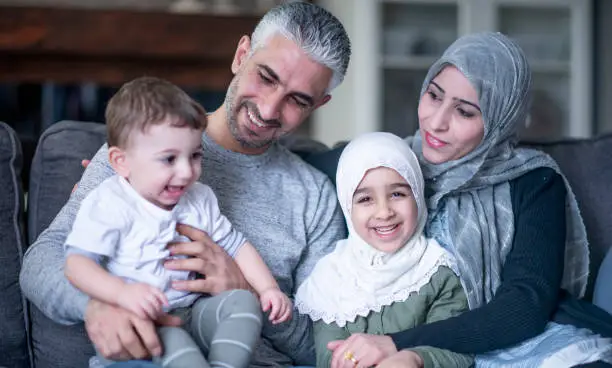 A beautiful Muslim family with two young children. They are seated on a couch in their living room. They are happy and smiling.